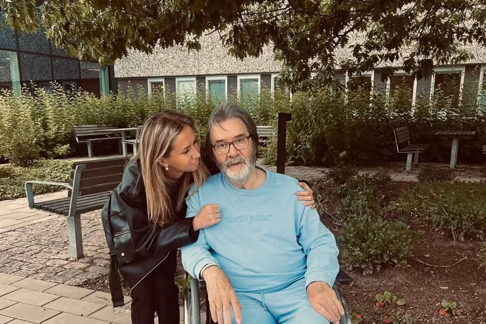 Horace Mason sitter i rullstol i en trädgård medan hans dotter Michelle lutar sig mot honom.